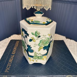 Może przedstawiać: Biały ceramiczny słoik o sześciokątnym kształcie z pasującą pokrywką. Słoik jest ozdobiony zielonymi i niebieskimi wzorami kwiatowymi i złotymi obramowaniami. Słoik stoi na niebieskiej książce.