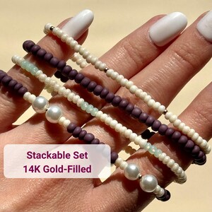May include: Stackable set of beaded bracelets in cream, purple, and light green. Some bracelets feature pearls. The text "Stackable Set 14K Gold-Filled" is visible on a white rectangle.