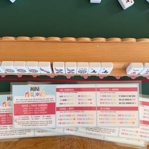 Peut inclure: Un support de mah-jong en bois contenant des tuiles blanches avec des symboles rouges, noirs et verts. En dessous, une carte d'instructions plastifi&eacute;e intitul&eacute;e Mini Mahjong pr&eacute;sente les r&egrave;gles et combinaisons en texte rouge, bleu et noir.