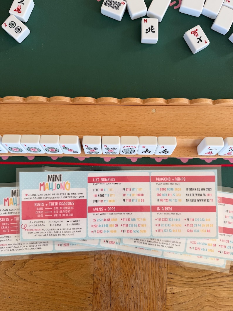 Peut inclure: Un support de mah-jong en bois contenant des tuiles blanches avec des symboles rouges, noirs et verts. En dessous, une carte d'instructions plastifi&eacute;e intitul&eacute;e Mini Mahjong pr&eacute;sente les r&egrave;gles et combinaisons en texte rouge, bleu et noir.