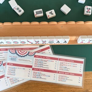 May include: A wooden mahjong tile rack holding white tiles with red, green, and black markings, positioned above a laminated score card titled Red, White + Mahj. The card lists game combinations like 1776, The 4th of Mahj, and Stars + Stripes.