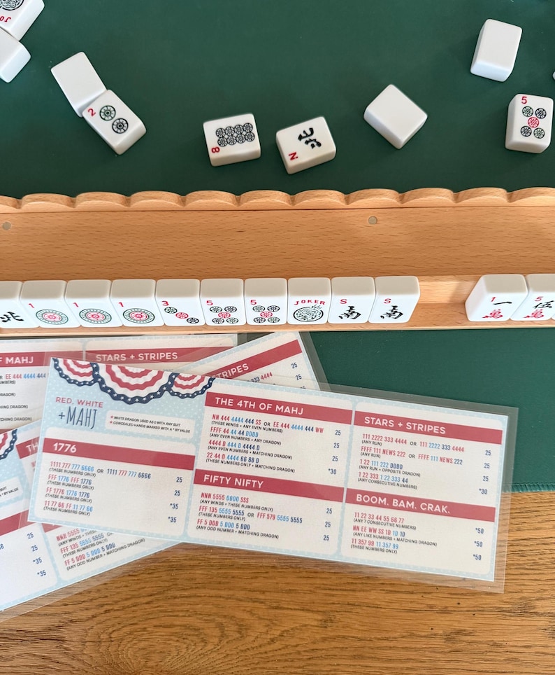 May include: A wooden mahjong tile rack holding white tiles with red, green, and black markings, positioned above a laminated score card titled Red, White + Mahj. The card lists game combinations like 1776, The 4th of Mahj, and Stars + Stripes.