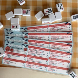 Pu&ograve; includere: Un set di tessere del Mahjong sparse attorno a diverse schede segnapunti plastificate Mahj-Oween. Le schede presentano grafiche a tema Halloween, tra cui zucche e pipistrelli, con testo che elenca mani di gioco come Curse of the Consecutives e Witch Way to the Joker.