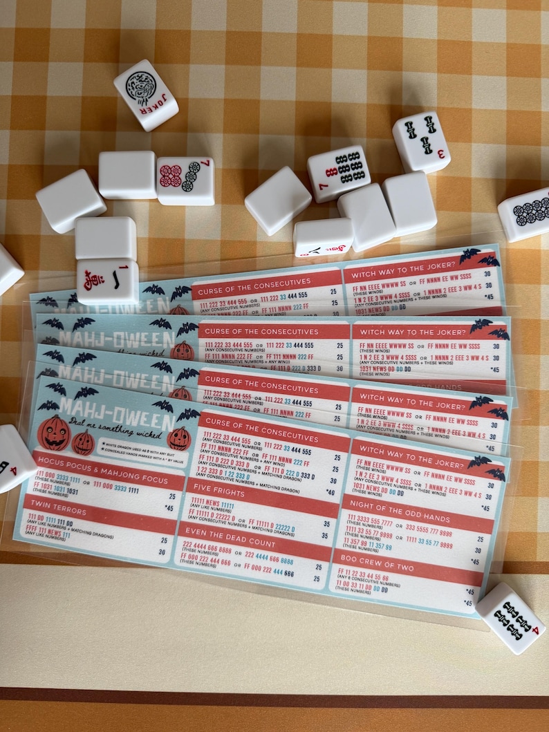 Pu&ograve; includere: Un set di tessere del Mahjong sparse attorno a diverse schede segnapunti plastificate Mahj-Oween. Le schede presentano grafiche a tema Halloween, tra cui zucche e pipistrelli, con testo che elenca mani di gioco come Curse of the Consecutives e Witch Way to the Joker.