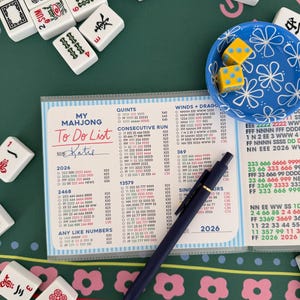 May include: A Mahjong To Do List, labelled "Katie", is visible on a clear plastic sheet. The sheet is on a green surface with Mahjong tiles and dice. A blue pen rests on the list, which includes numbers and Mahjong tile combinations.