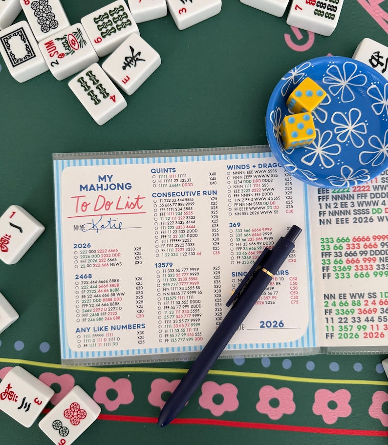 May include: A Mahjong To Do List, labelled "Katie", is visible on a clear plastic sheet. The sheet is on a green surface with Mahjong tiles and dice. A blue pen rests on the list, which includes numbers and Mahjong tile combinations.