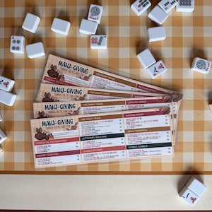Pu&ograve; includere: Una pila di quattro segnapunti Mahj-Giving a tema Ringraziamento, con testo che include Mahj-Giving, The Gravy Train e Autumn Winds, circondate da tessere del Mahjong bianche sparse su una superficie a scacchi beige e bianca.