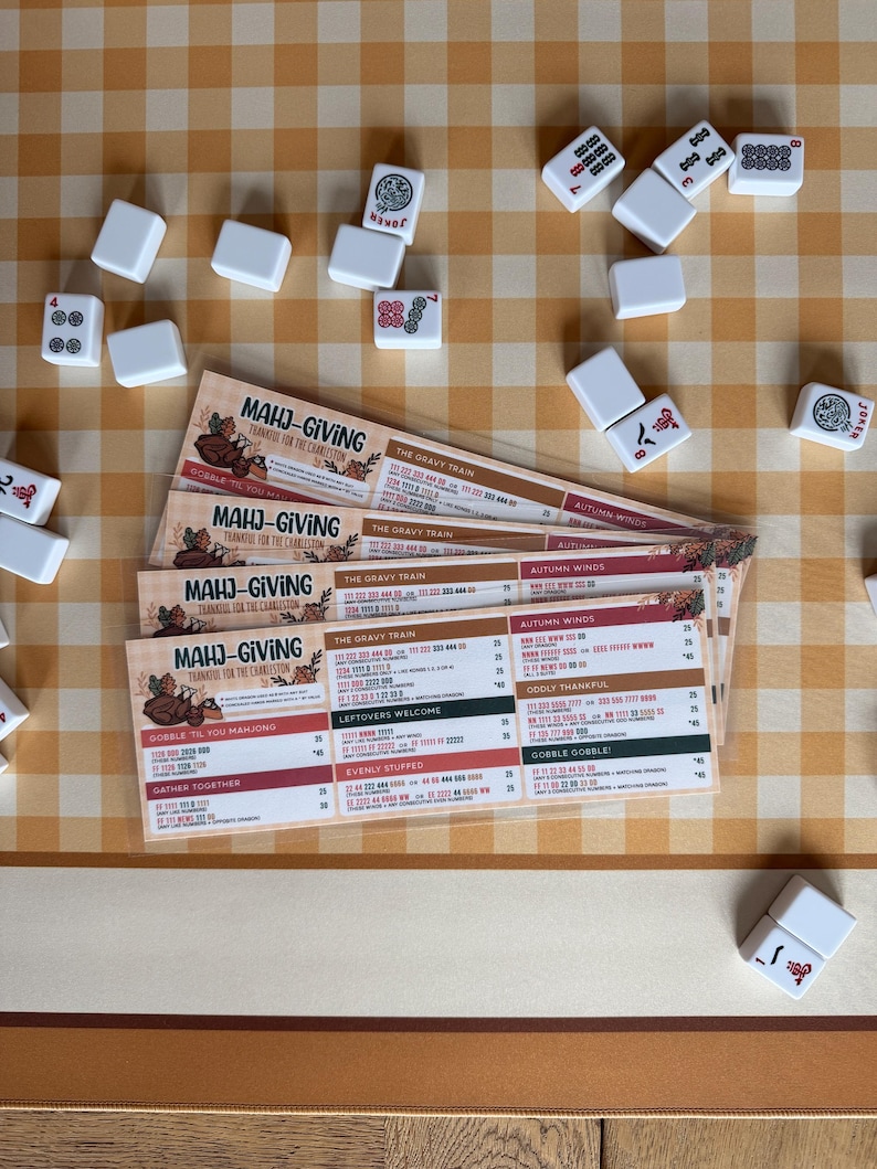 Pu&ograve; includere: Una pila di quattro segnapunti Mahj-Giving a tema Ringraziamento, con testo che include Mahj-Giving, The Gravy Train e Autumn Winds, circondate da tessere del Mahjong bianche sparse su una superficie a scacchi beige e bianca.