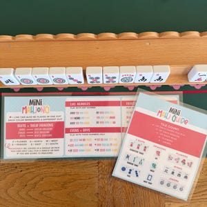 Peut inclure: Un support &agrave; tuiles de Mahjong en bois contenant des tuiles blanches avec divers symboles et chiffres, accompagn&eacute; de deux cartes d'instructions plastifi&eacute;es intitul&eacute;es Mini Mahjong et Mini Mahj Guide, d&eacute;taillant les combinaisons de jeu.