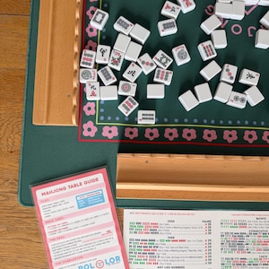 May include: A Mahjong set with white tiles scattered on a green playing surface. The set includes a wooden table, a pink and white Mahjong Table Guide, and a wooden tray. The tiles have various symbols and characters.