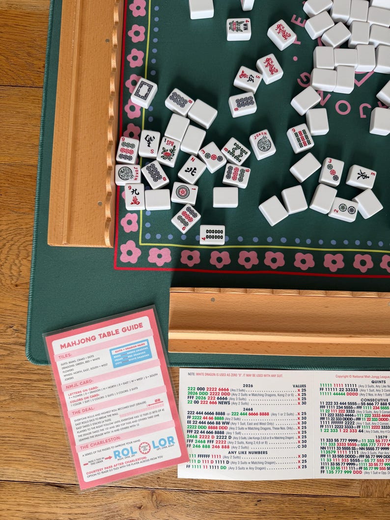 May include: A Mahjong set with white tiles scattered on a green playing surface. The set includes a wooden table, a pink and white Mahjong Table Guide, and a wooden tray. The tiles have various symbols and characters.