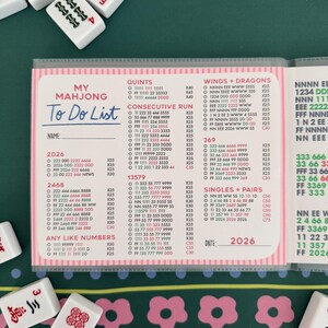 K&ouml;nnte beinhalten: Ein Mahjong To Do List Notizblock mit rosa Akzenten, der Zahlenkombinationen und Spielstrategien zeigt. Der Notizblock enth&auml;lt das Jahr 2026 und ist von Mahjong-Steinen umgeben.