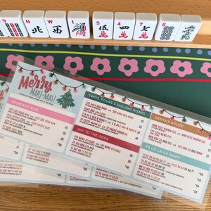 Pu&ograve; includere: Una scheda segnapunti plastificata per Mah Jongg intitolata Merry Mahj-Mas con grafiche a tema natalizio, tra cui luci a stringa e un albero di Natale. La scheda elenca varie combinazioni di mani come Reindeer Run e Santa's Route con i punti.