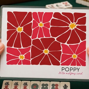 May include: A card with a floral design featuring red and pink flowers with yellow centers. The word "POPPY" is printed in black, with "2026 mahjong card" in pink below. The card is held in front of a green surface with mahjong tiles.