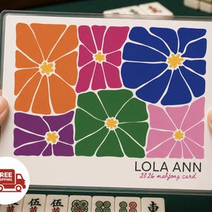 May include: A rectangular mahjong card encased in a clear plastic sleeve, featuring a grid of six stylized flowers in orange, magenta, blue, purple, green, and light pink. The text reads LOLA ANN 2026 mahjong card in black and pink lettering.