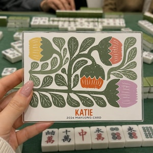Könnte beinhalten: Eine transparente Plastikkarte mit einem floralen Design mit gelben, orangefarbenen und lila Blumen und grünen Blättern. Auf der Karte steht "KATIE 2026 MAHJONG CARD". Die Karte wird vor einer grünen Oberfläche mit Mahjong-Steinen gehalten.