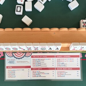 May include: A wooden mahjong tile rack holding white tiles with red, green, and black symbols, including a Joker tile. Below the rack is a laminated Red, White + Mahj score card featuring game rules and combinations like 1776, Fifty Nifty, and Stars + Stripes.