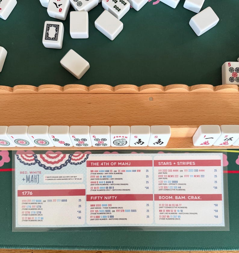 May include: A wooden mahjong tile rack holding white tiles with red, green, and black symbols, including a Joker tile. Below the rack is a laminated Red, White + Mahj score card featuring game rules and combinations like 1776, Fifty Nifty, and Stars + Stripes.