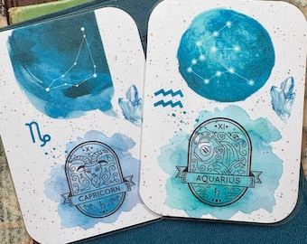 May include: Two white cards with rounded corners, featuring watercolor designs of the Capricorn and Aquarius zodiac signs. Each card has a blue watercolor background, constellation illustrations, and crystal accents. The cards are on top of books.