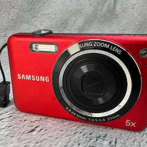 May include: A bright red Samsung digital camera featuring a prominent zoom lens with 4.9-24.5mm 1:3.5-5.9 27mm specifications printed on the black ring. The device includes a built-in flash, a 5x optical zoom indicator, and a black wrist strap.