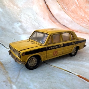 Puede incluir: Un coche de juguete amarillo con rayas negras y la palabra "милиция" impresa en el lateral. El coche de juguete tiene un emblema rojo. Es un modelo en miniatura.