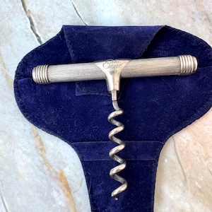 以下が含まれることがあります： リブ付きハンドルとスパイラルワームが付いた銀色のコルクスクリュー。濃い青色のベルベットポーチの上に置かれています。「Cliente」の文字が刻印されています。ポーチは開いており、コルクスクリューが見えています。