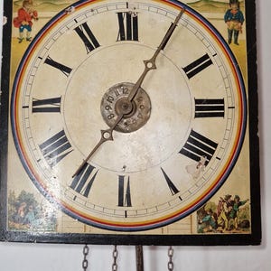 May include: Vintage wall clock with a painted face and a black frame. The clock face has Roman numerals, a rainbow border, and a pastoral scene at the top. The clock has decorative figures on each side and a pendulum with chains.