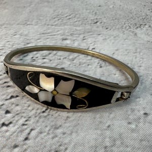 Könnte beinhalten: Ein aufklappbarer Metallarmreif mit einer schwarzen Einlage, die mit einem weißen Perlmutt-Blumenmotiv verziert ist. Das Armband hat ein poliertes silberfarbenes Finish und einen sicheren Verschlussmechanismus an der Seite mit einem floralen Design.