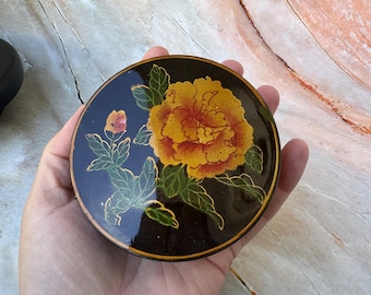 May include: A round, black decorative box with a vibrant yellow and orange floral design. The flower is the focal point, with green leaves and gold accents. The box is held in a hand.