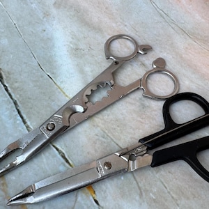 May include: Two pairs of metal tools on a light-colored surface. One tool is a silver multi-tool with a bottle opener and pliers. The other is a pair of scissors with black handles and silver blades. Fishing tools.