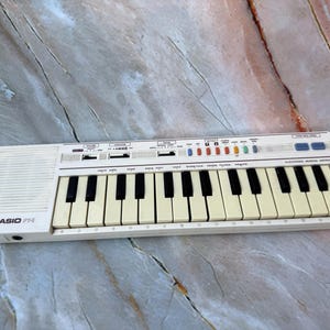 Puede incluir: Un instrumento musical electrónico Casio PT-14 blanco con teclas negras y marfil. El teclado tiene un altavoz, botones de control y una serie de botones de colores. El texto "CASIO PT-14" está impreso en el lado izquierdo.