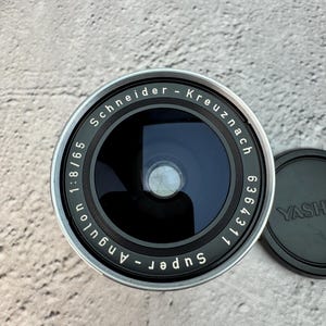 May include: A Schneider-Kreuznach Super-Angulon 1:8/65 camera lens with a black front element and silver metal housing. The lens features white text around the glass, including the serial number 6364311. A black lens cap with YASHICA text is nearby.