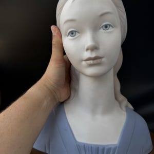Puede incluir: Busto de porcelana de una mujer con piel pálida, ojos azules y cabello claro. Viste un vestido azul con escote cuadrado. El busto está montado sobre una base de madera.
