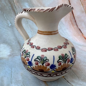 Könnte beinhalten: Eine cremefarbene Keramikkrug mit Henkel und Ausguss. Der Krug ist mit braunen, roten, blauen und grünen Blumenmustern verziert. Ein braunes Band umschließt die Mitte, und der Rand hat einen dekorativen Rand.