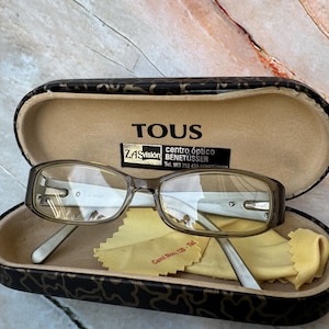 Könnte beinhalten: Brille in einem schwarz-braun gemusterten Etui mit dem Markennamen TOUS. Die Brille hat einen hellbraunen Rahmen und klare Gläser. Ein gelbes Reinigungstuch befindet sich im Etui.