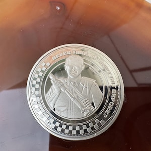 May include: A silver coin featuring a man in uniform holding a weapon. The coin has the text "NEVER IN A SHORT CUT BEFORE?" around the edge. The coin has a checkered pattern around the edge.