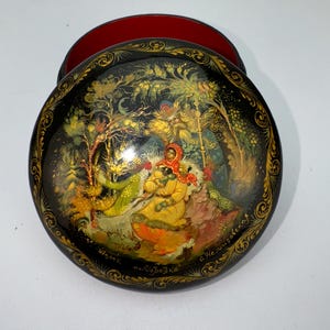 Op de afbeelding: Een ronde zwarte lakdoos met een rode binnenkant, voorzien van een handgeschilderde scène van een figuur in een gele jas en rode hoofddoek in een bosrijke omgeving. Het deksel bevat gouden filigraan randdetails en cyrillische tekst onderaan.