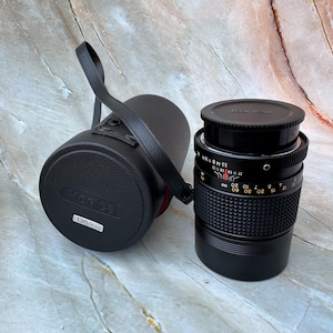 May include: A black Konica camera lens with a matching case. The lens has a black cap and a series of measurement markings. The case has a black strap and the Konica logo. The lens is a 135/3.5 model.