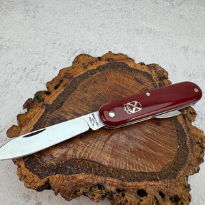 以下が含まれることがあります： 銀色の刃と装飾的なエンブレムが付いた、バーガンディ色のポケットナイフ。 ナイフは、自然な質感の木片の上に置かれています。 ナイフの長さは約10cmです。