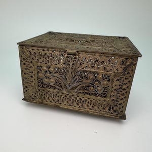 Könnte beinhalten: Eine antike Messing-Filigran-Box mit Scharnierdeckel. Die Box weist aufwendige Durchbruchmuster und florale Designs auf. Das Metall hat ein angelaufenes, gealtertes Aussehen, was auf Vintage hindeutet. Die Box ist rechteckig und vermutlich eine Schmuck- oder Schmuckschatulle.