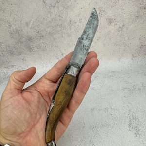 Puede incluir: Una navaja plegable vintage con una hoja plateada desgastada y un mango curvo de madera. El mango tiene un tono marrón cálido con vetas de madera visibles. La navaja está abierta, mostrando toda la longitud de la hoja. Los componentes metálicos muestran signos de edad.