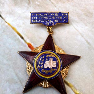 Könnte beinhalten: Eine sternförmige Vintage-Medaille mit weinroter Emaille und goldfarbenen Akzenten. Die Medaille hat einen blauen rechteckigen Balken mit Text und dem Jahr 1970. Die Mitte hat ein blau-gelbes kreisförmiges Emblem mit einem Gebäudedesign.