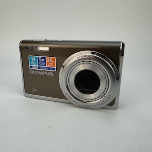 Op de afbeelding: Een bruine Olympus FE digitale camera met een zilveren lens en flitser. De camera heeft een sticker met de tekst "14 MEGA PIXELS", "5x OPTICAL ZOOM" en "24 WIDE". De lens heeft de tekst "OLYMPUS LENS" en "5x WIDE OPTICAL ZOOM".