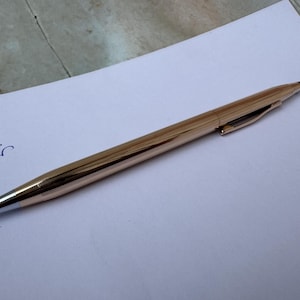 Könnte beinhalten: Ein goldfarbener Stift mit silberner Spitze und schwarzem Oberteil, der auf einem weißen Blatt Papier liegt. Der Stift hat einen Clip an der Seite. Das Wort "Cross" ist mit blauer Tinte auf das Papier geschrieben.