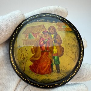 May include: A round black lacquer box featuring a hand-painted scene of two figures in traditional winter clothing standing in a field with small structures in the background. The lid has a gold-painted decorative border with crescent shapes.