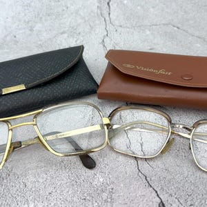 Puede incluir: Dos pares de gafas con monturas doradas y lentes transparentes, junto con dos estuches. Un estuche es negro con detalles dorados y el nombre "Dakora". El otro estuche es marrón con el nombre "Visionfast".