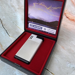 May include: A silver and black lighter with a textured surface, presented in a black box with a red velvet interior. The box features a landscape painting and the text "KAWEE".