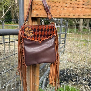 May include: Brown leather crossbody bag with a sunflower design and diamond pattern. The bag features long fringe and a brown leather strap. The bag is hanging on a metal fence post.