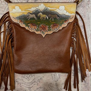 May include: Brown leather crossbody bag with a painted landscape scene of horses running in front of mountains. The bag has a flap with a floral design and long fringe on the sides. The bag has a shoulder strap.