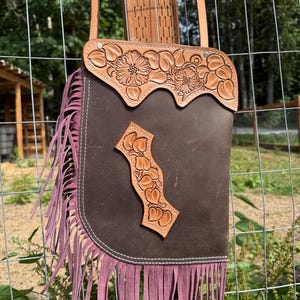 May include: A brown leather crossbody bag with a floral embossed design and a fringe detail. The bag has a light brown strap and a decorative floral design on the flap. The bottom of the bag features a mauve-coloured fringe.